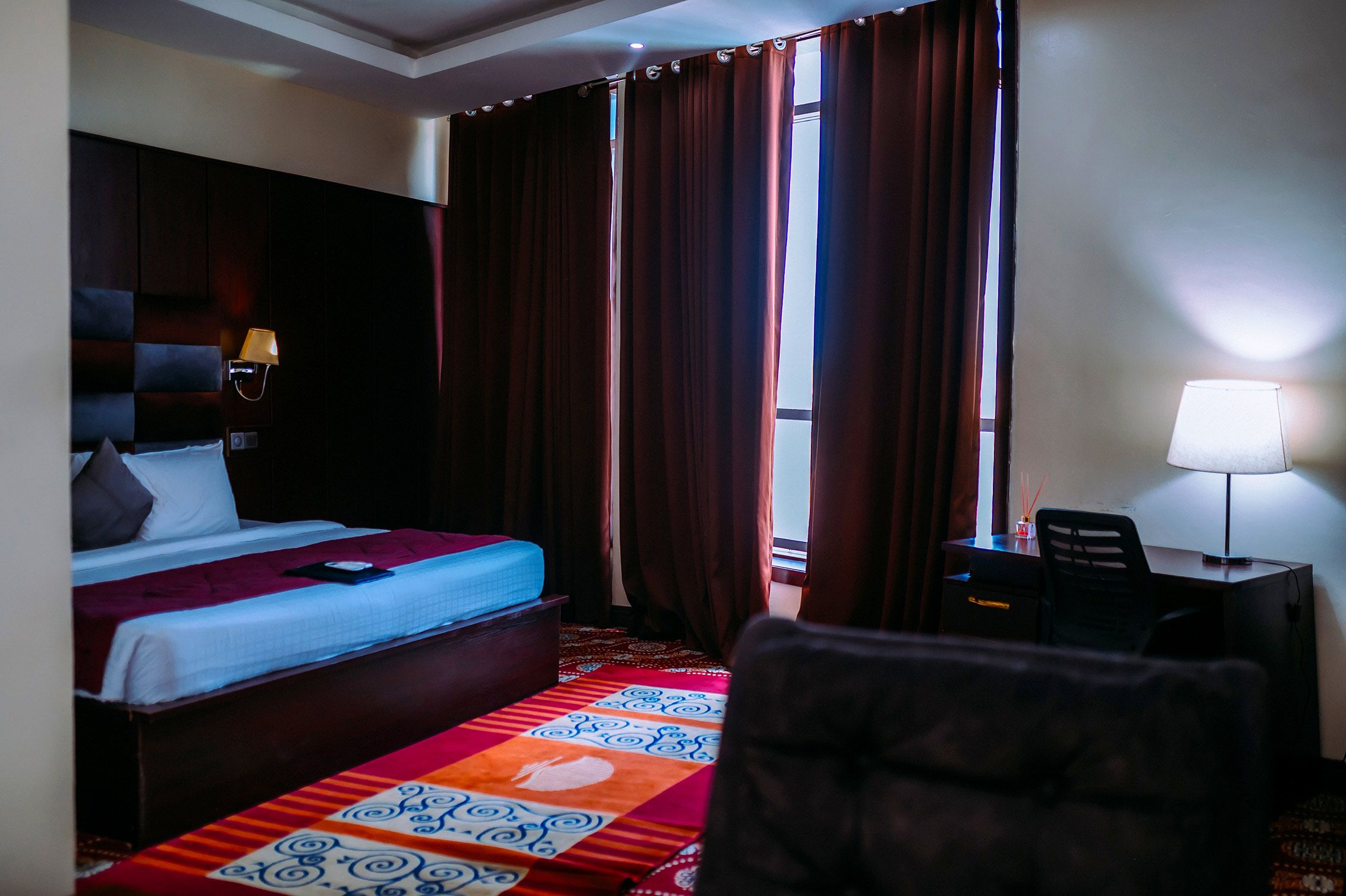 A hotel room with a bed, a desk, a chair, a lamp, and a window with curtains.