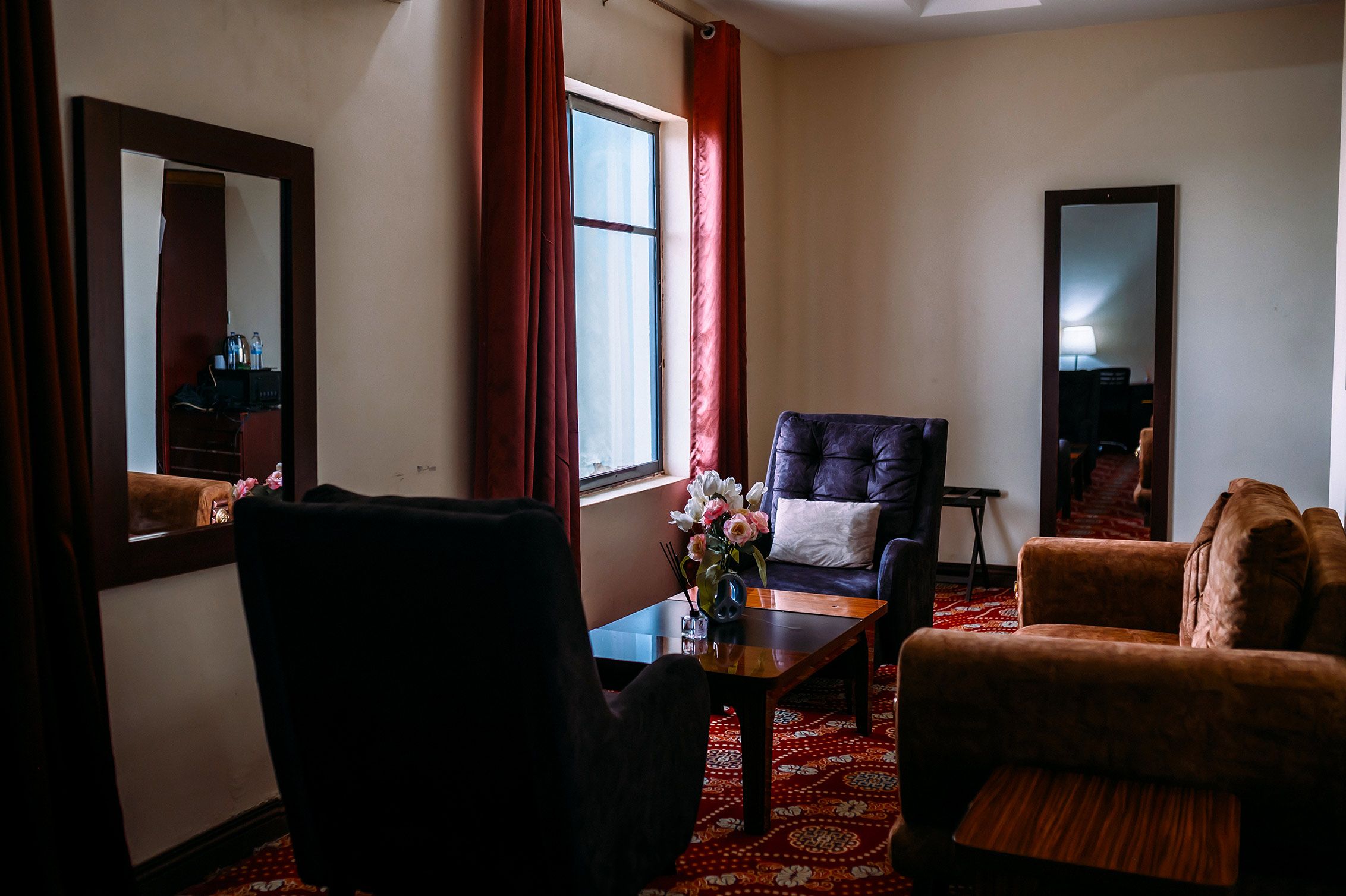 Una habitación con un sillón oscuro, un sofá, una mesita con un jarrón de flores y un espejo grande en la pared.