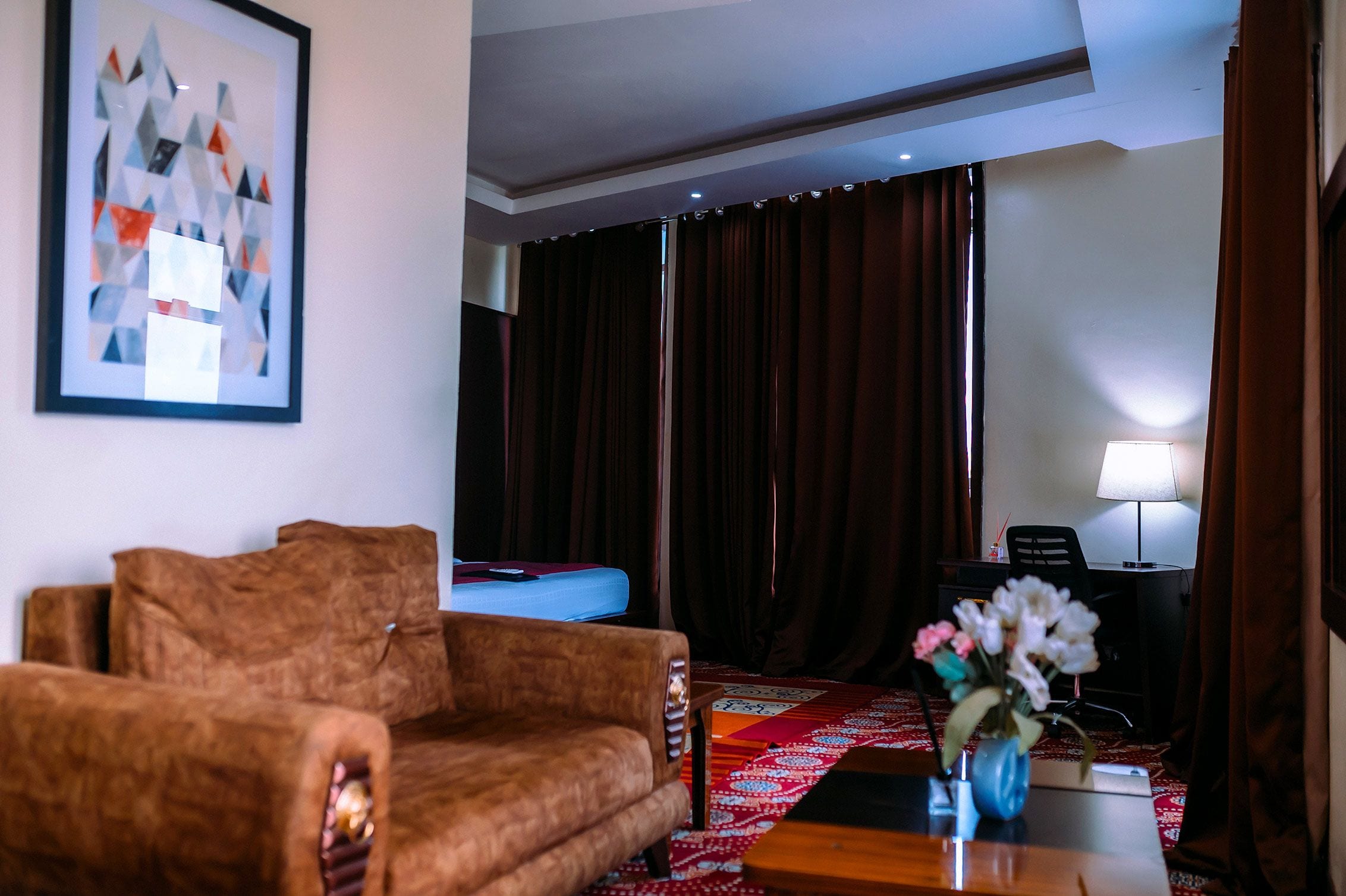 A hotel room with a brown sofa, a bed with a blue headboard, a red patterned rug, a desk with a lamp, and a framed abstract painting on the wall.