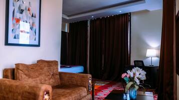 A hotel room with a brown sofa, a bed with a blue headboard, a red patterned rug, a desk with a lamp, and a framed abstract painting on the wall.