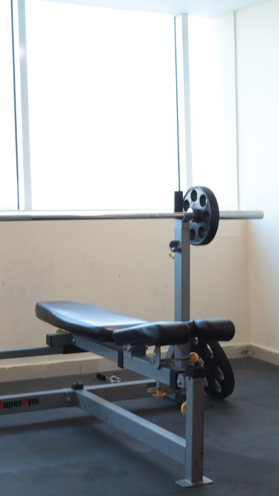 A bench with a weight stack and a barbell in a room with a window and a plain wall.