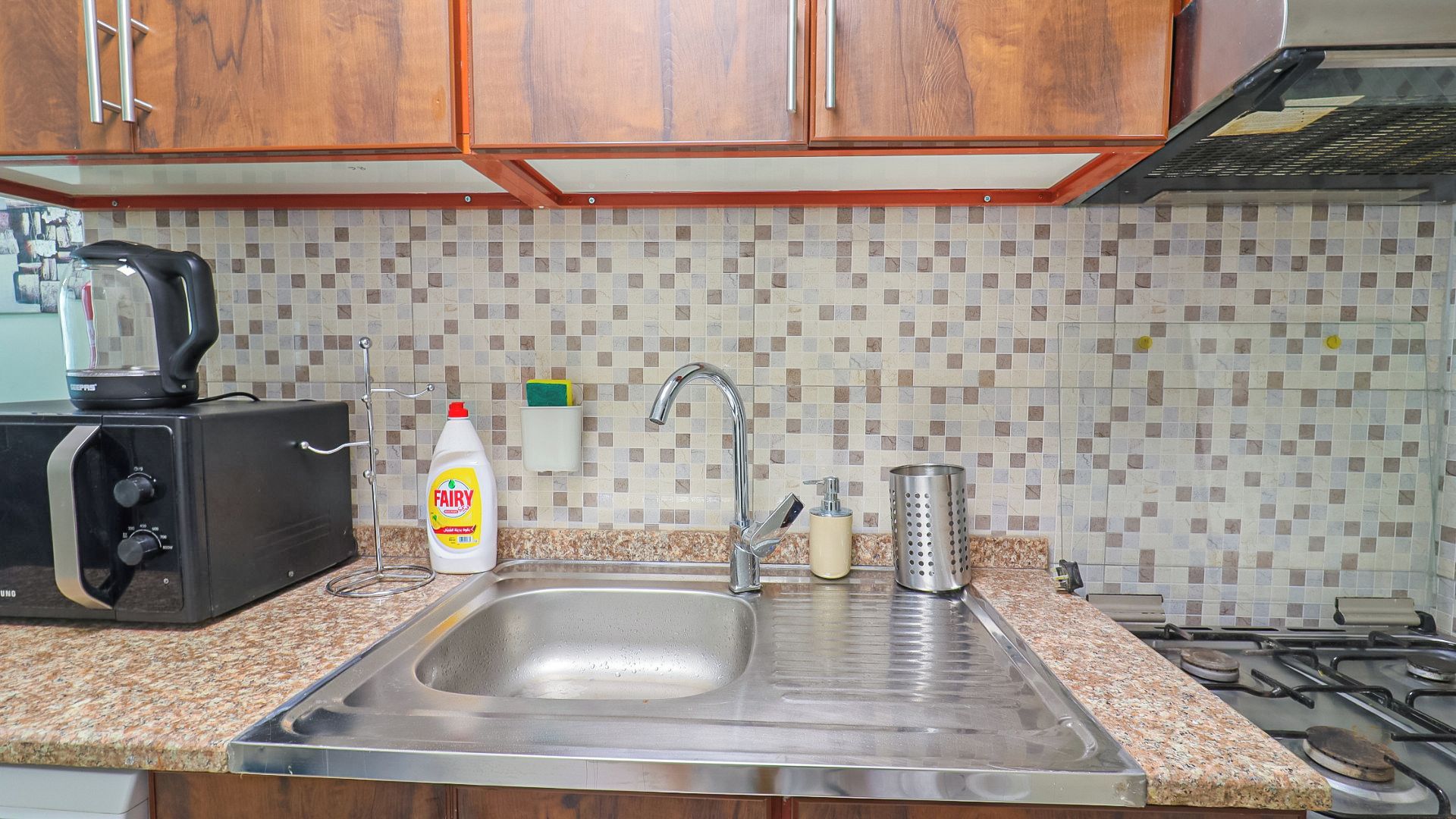A kitchen with a stainless steel sink, a kettle, a bottle of Fairy dish soap, and a grater.