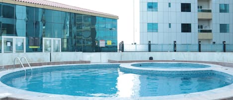 A swimming pool with a tiled edge, surrounded by a low wall and a building with reflective glass windows in the background.