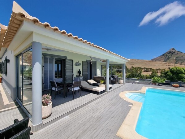 A modern outdoor pool area with a covered seating area, a clear blue sky, and a mountainous backdrop.