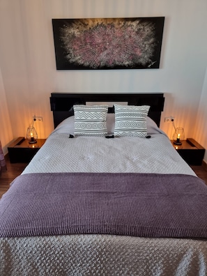 Un lit bien fait, agrémenté d’oreillers à motifs et d’une couverture texturée, flanqué de tables de chevet avec des lampes, dans une pièce où un grand tableau abstrait orne le mur.