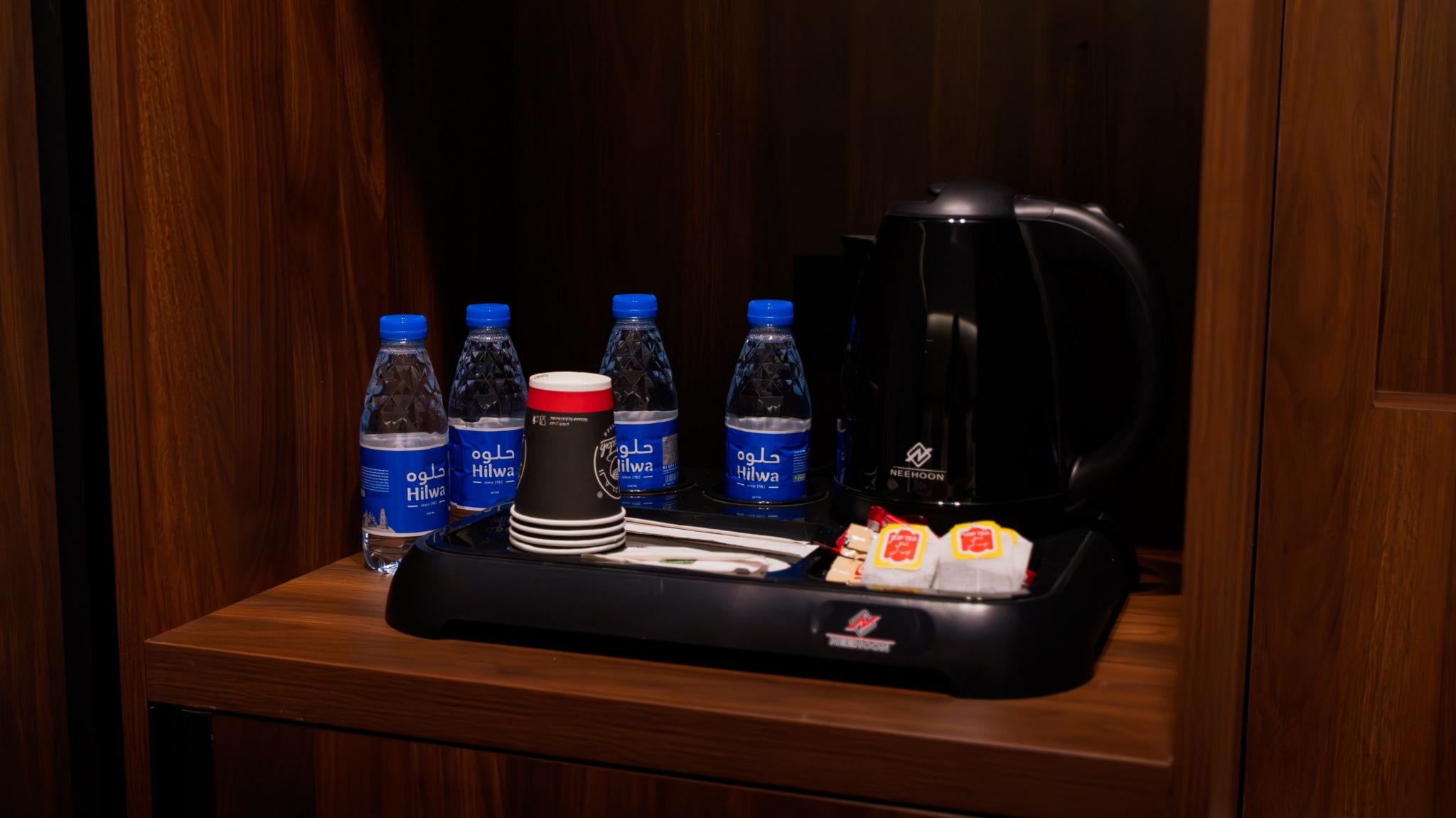 A hotel room with a wooden table, a black tray with a coffee maker, water bottles, a red cup, and a stack of white plates.
