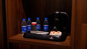 Habitación de hotel con una mesa de madera, una bandeja negra que contiene una cafetera, botellas de agua, una taza roja y una pila de platos blancos.