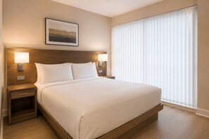 Ein Hotelzimmer mit einem großen Bett, zwei Nachttischlampen, einem Nachttisch, einem gerahmten Bild an der Wand und einem Fenster mit Jalousien.