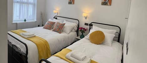 A hotel room with two single beds, each with a yellow throw blanket and white bedding. There are two wall-mounted lamps and two abstract paintings on the walls.
