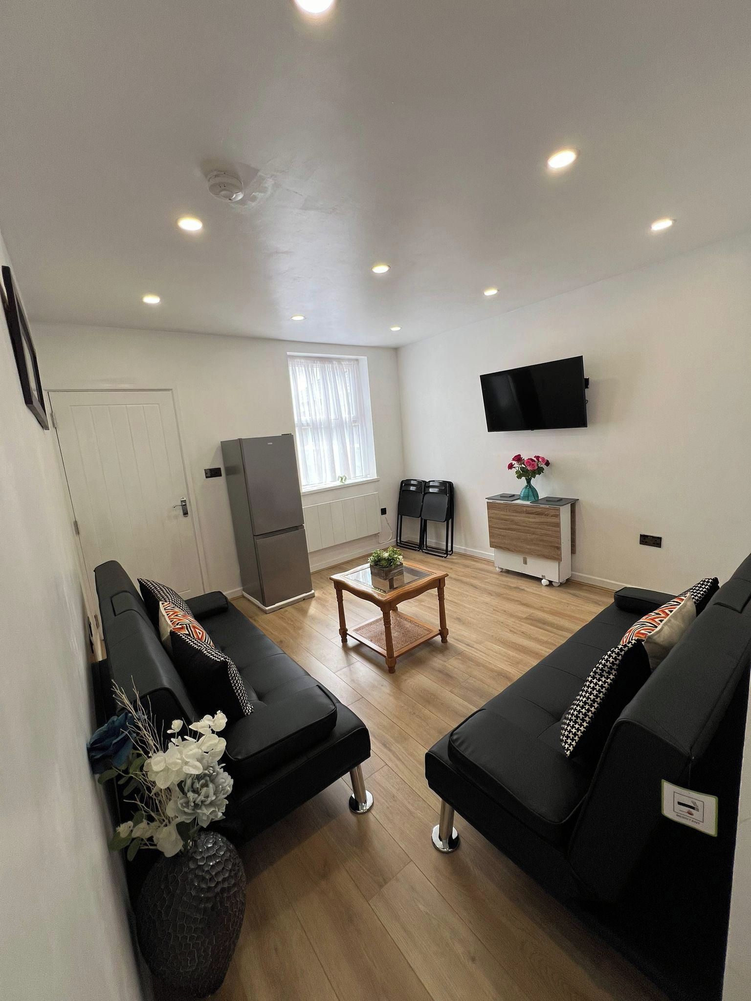 A living room with two black sofas, a coffee table, a TV on the wall, a refrigerator, and a window with curtains.