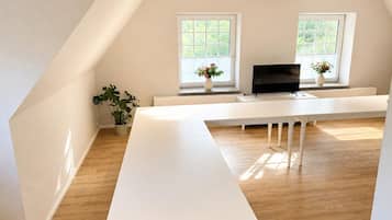 A modern, minimalist room with a white desk, two windows with blinds, a flat-screen TV, and a potted plant.
