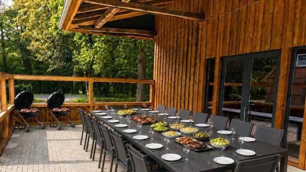 An outdoor dining area with a long table set for a meal, surrounded by chairs, and a grill in the background.