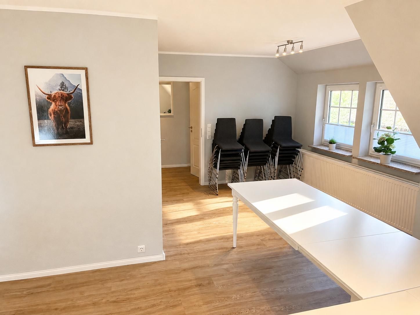 Ein Zimmer mit einem weißen Tisch, schwarzen Stühlen, einem gerahmten Bild an der Wand und einem Fenster mit Pflanzen.