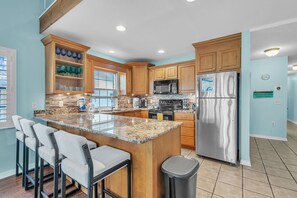 A modern kitchen with granite countertops, wooden cabinets, a stainless steel refrigerator, and a bar area with chairs. - On the Intracoastal | Pool | Fishing Pier | Walk to Beach | Indian Sands B3 (Indian Shores)