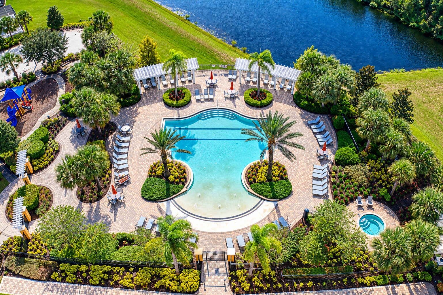 An aerial view of a resort with a central swimming pool, surrounded by lounge areas and palm trees.
