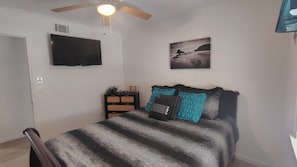 Une chambre à coucher avec un lit, un téléviseur fixé au mur, un ventilateur de plafond et un tableau encadré.