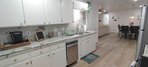 A kitchen with white cabinets, a dishwasher, a sink, and a dining area with a table and chairs. - Brand New Remodel Off I-30 Mins to Buc-ee's w/ Ample Parking (Benton)