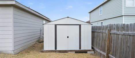 A small shed with a white door and a black handle, located in a backyard with dry grass and a wooden fence.