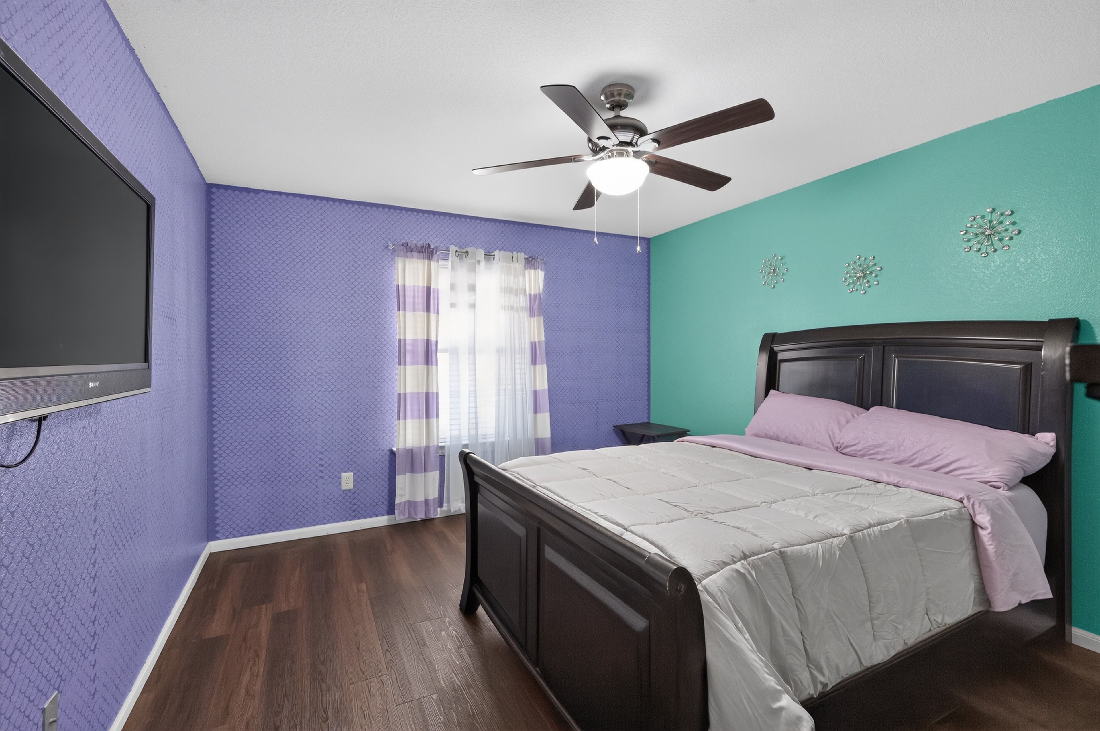 A bedroom with a bed, a ceiling fan, a window with curtains, and a wall-mounted TV.