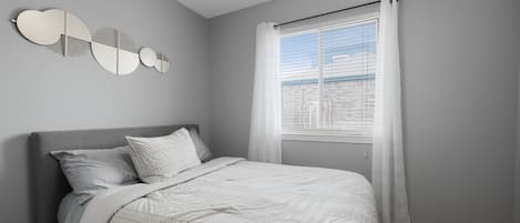 A bedroom with a bed, a window with curtains, and a wall-mounted light fixture.