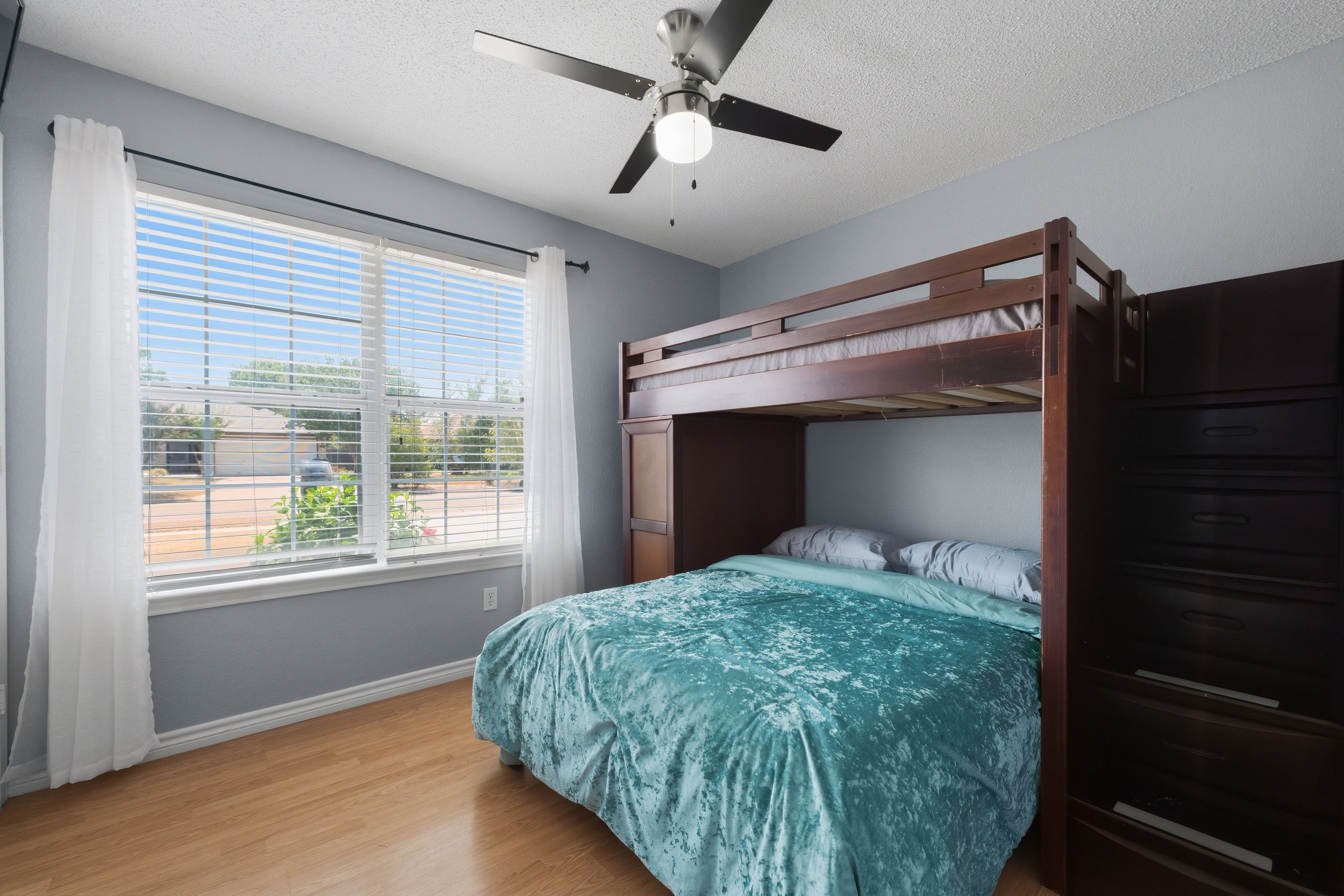A room with a bunk bed, a ceiling fan, a window with blinds, and a view of the outside.