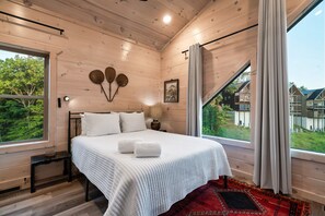 A bedroom with a bed, bedside table, lamp, and a view of greenery outside. - Smokiedoki Lodge 3 Bedroom Cabin (Pigeon Forge)