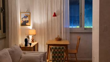 A living room with a sofa, a table with a chair, a lamp, a painting on the wall, and a window with curtains.