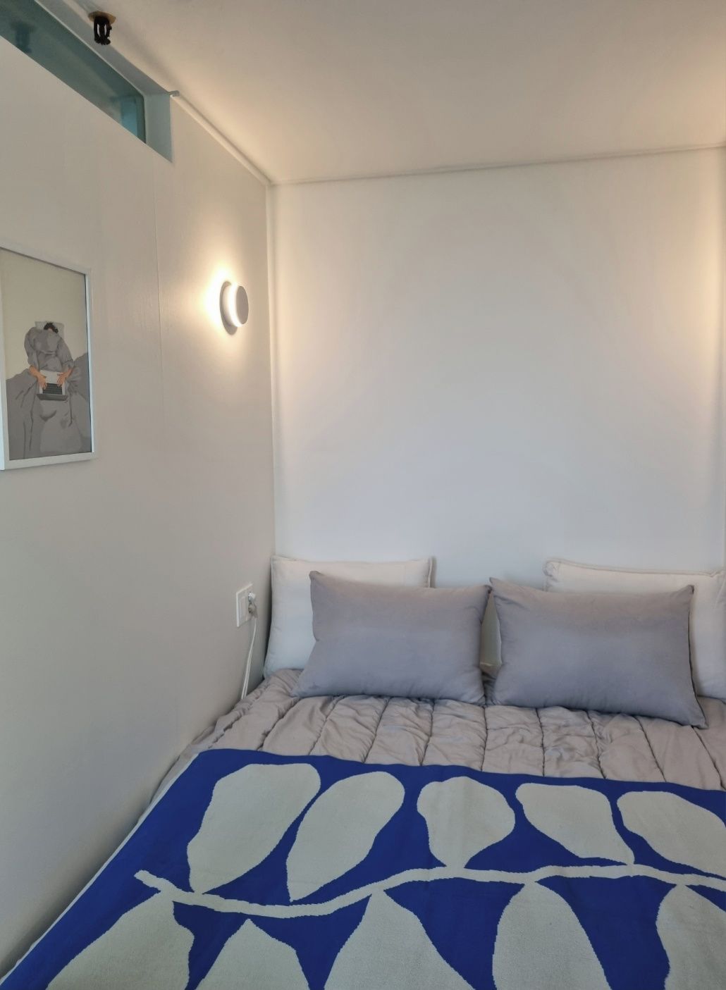 A bedroom with a bed, two pillows, a wall-mounted light, and a framed picture.