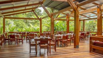 An indoor dining area with wooden tables and chairs, surrounded by a wooden structure with a metal roof, and a view of lush greenery outside.
