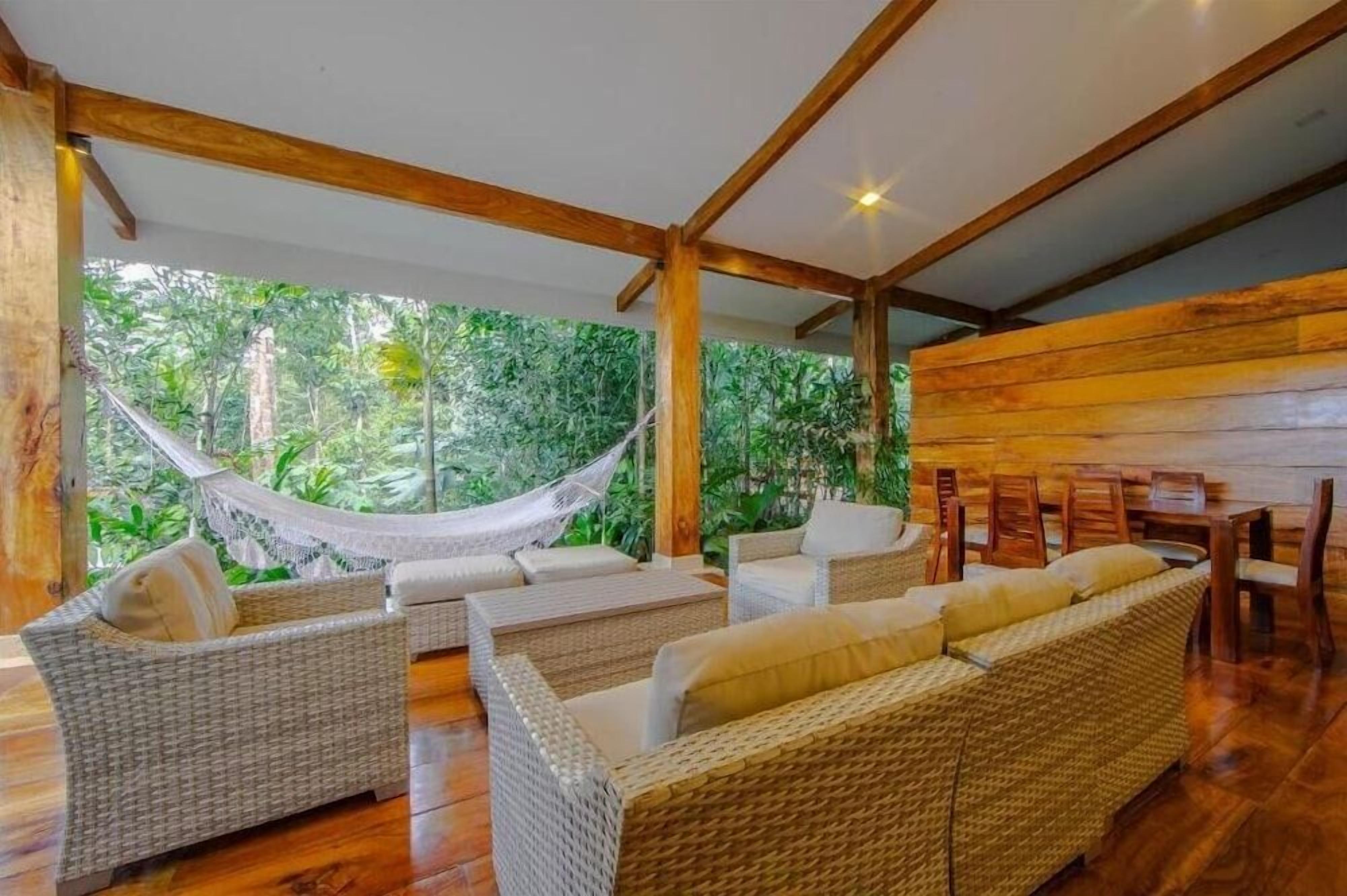 A room with wicker furniture, a hammock, and a view of lush greenery.