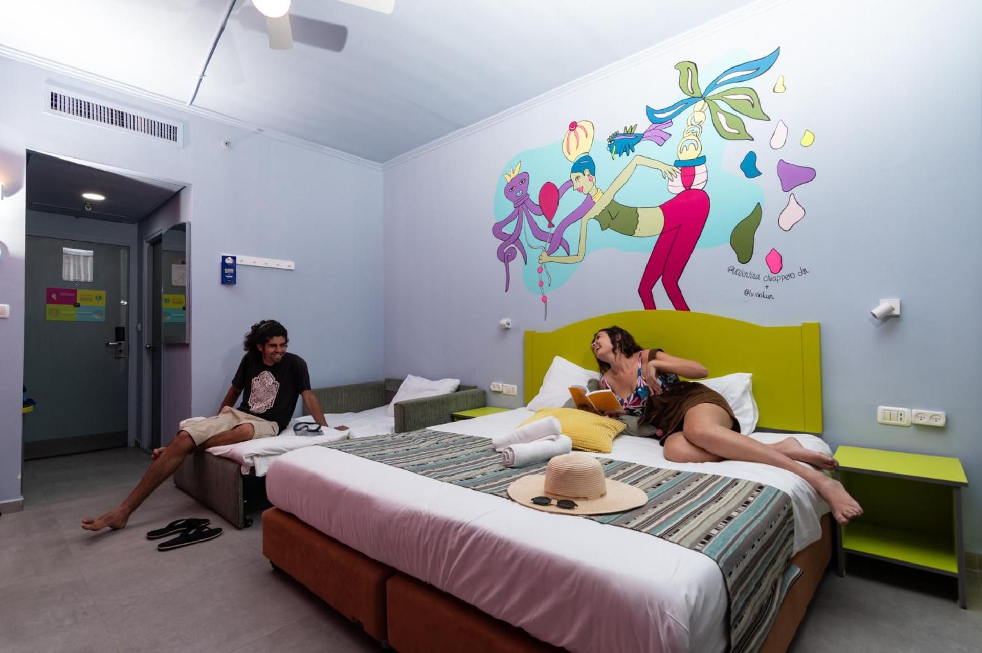 A hotel room with a bed, a bedside table, a wall with a colorful mural, and a door.