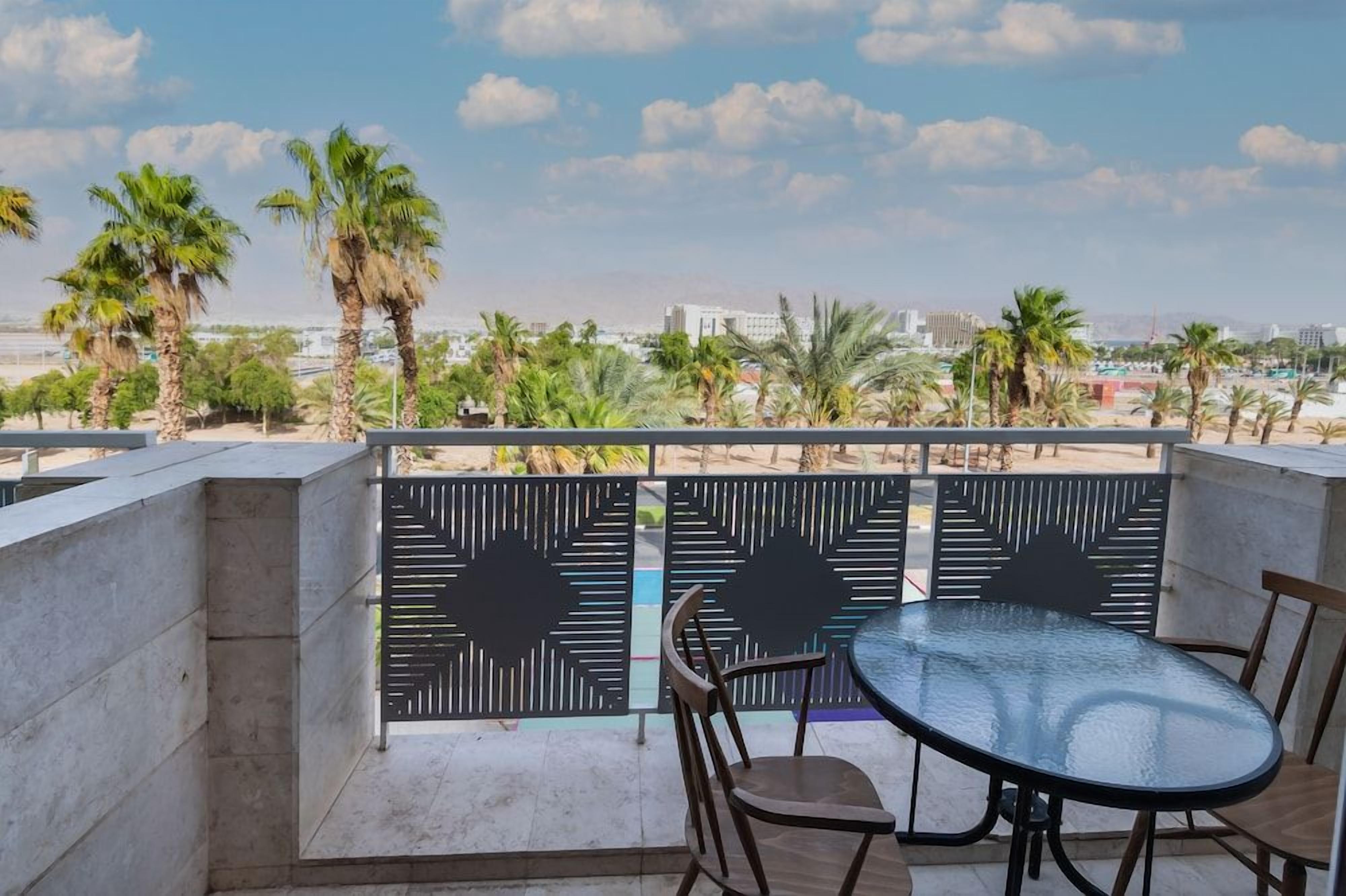 A balcony with a glass table and chairs, overlooking palm trees and a cityscape.
