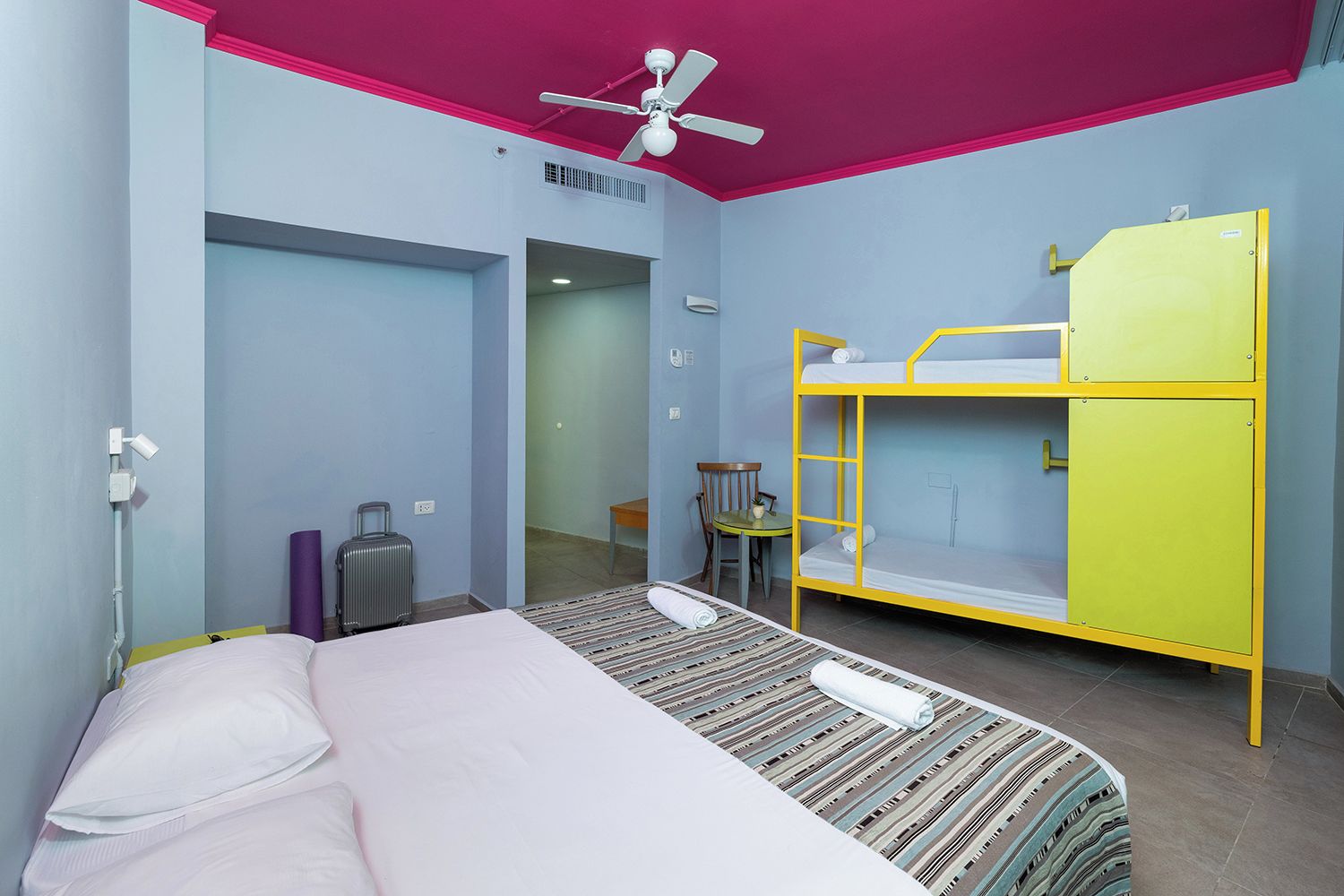 Une chambre avec un lit superposé, un lit simple, un ventilateur de plafond, une table avec une chaise et une valise.