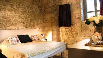 A bedroom with a stone wall, a bed with a white headboard, a bedside table with a lamp, and a window with curtains.