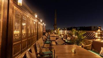 Een buitenruimte met houten tafels en stoelen, met uitzicht op een hoge toren in de verte, en een houten hek dat de ruimte omheint.
