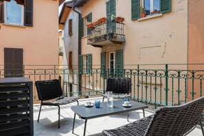 An outdoor seating area with a table and chairs, a balcony with potted plants, and a building with green shutters. - Casa Noce by J&CO Homes (CERNOBBIO)