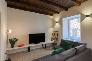 Ein modernes Wohnzimmer mit einem Flachbildfernseher auf einer weißen Konsole, einem grauen Sofa mit grünen Kissen, einer Topfpflanze und einem gerahmten Bild an der Wand.