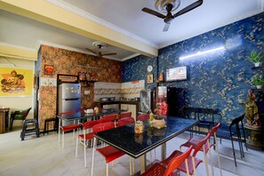 Un comedor con una mesa negra y sillas rojas, un ventilador de techo, y una cocina con refrigerador y estufa.