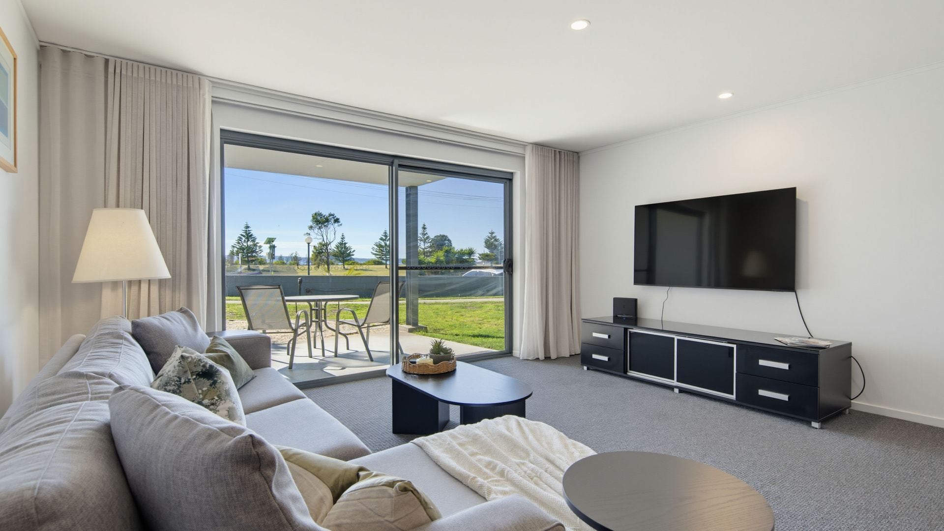A modern hotel room with a large sofa, a flat-screen TV, a dining area with a table and chairs, and a sliding glass door leading to a view of a golf course.