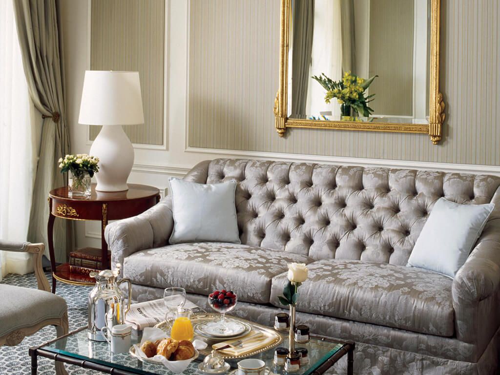 A luxurious living room with a tufted sofa, a glass coffee table with breakfast items, a side table with a lamp, and a large mirror.
