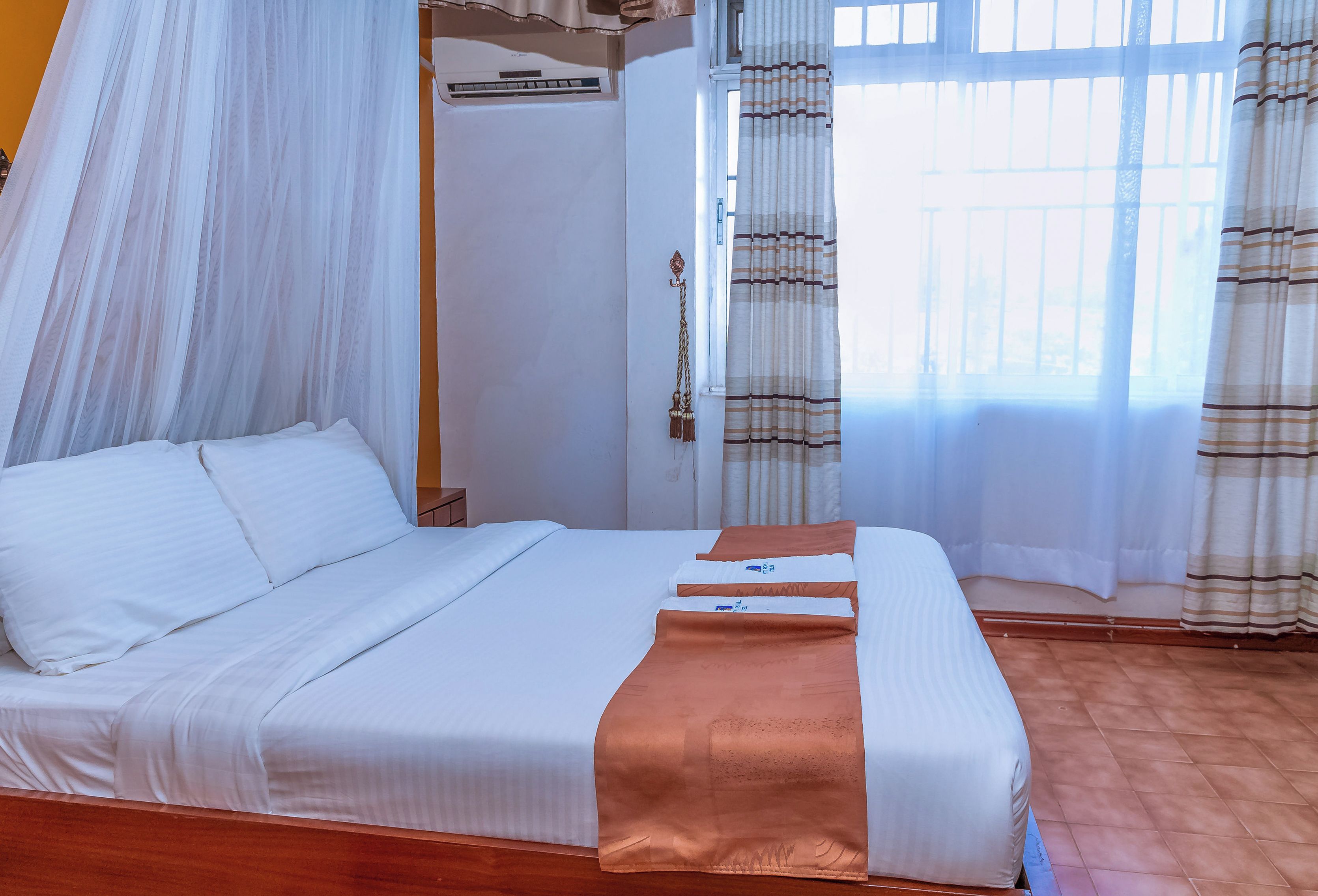 Quarto com cama, mesa de cabeceira, janela com cortinas e aparelho de ar condicionado.