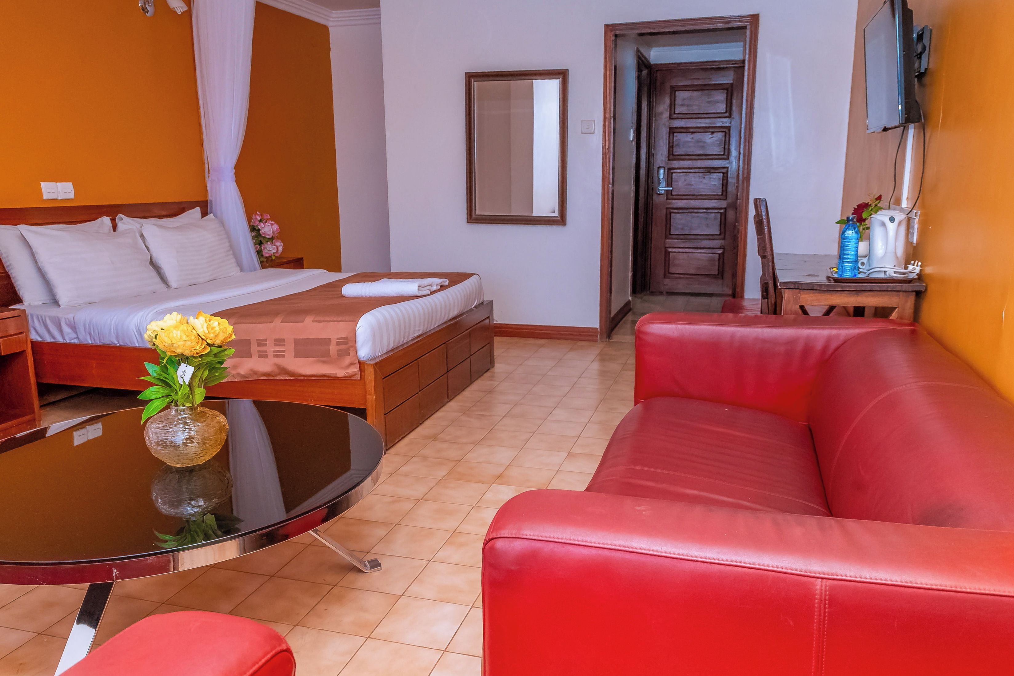 A hotel room with a bed, a red leather sofa, a small table with a vase of flowers, and a mirror on the wall.