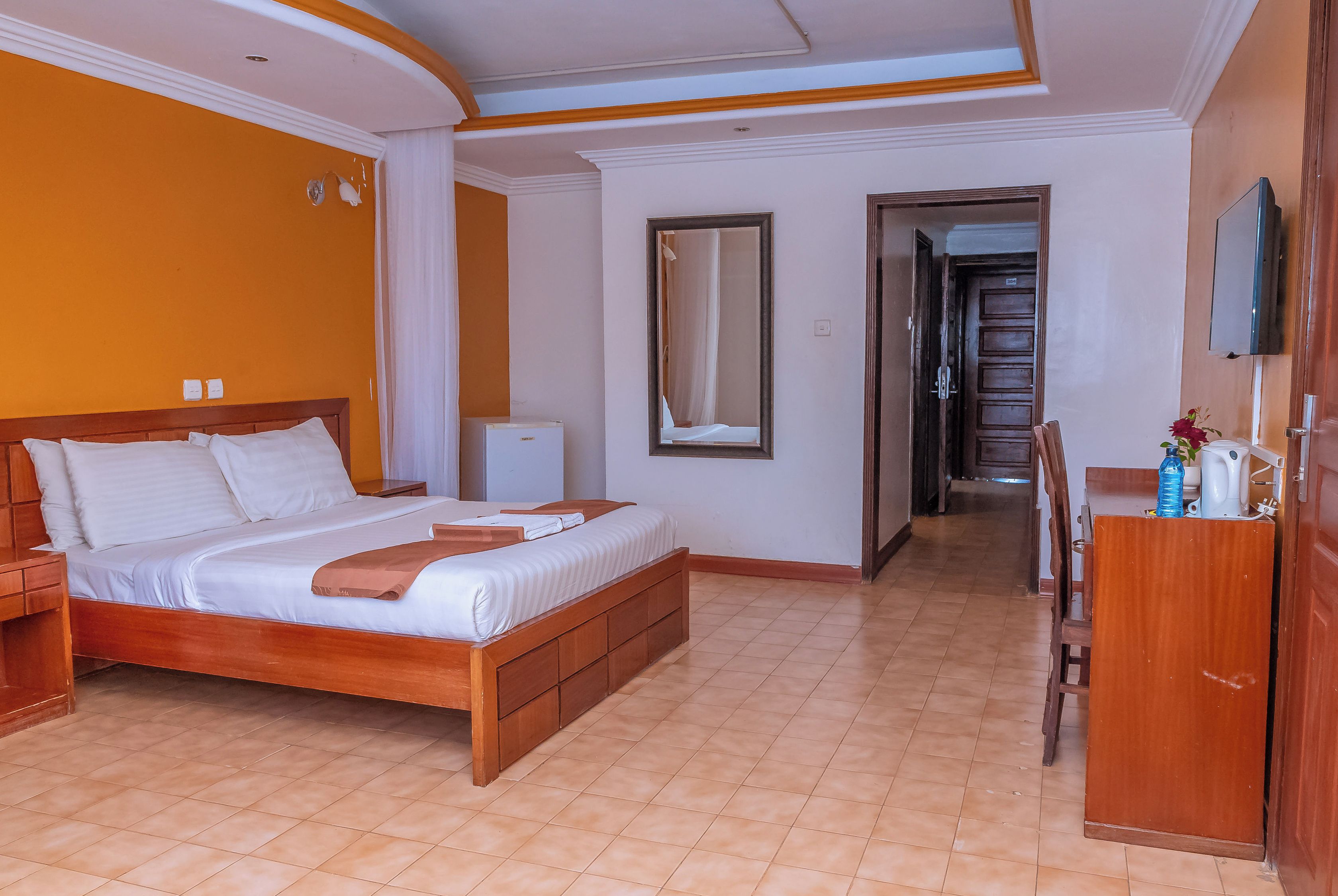 Une chambre d’hôtel avec un lit en bois, une table de chevet, un miroir, une armoire et un téléviseur fixé au mur.