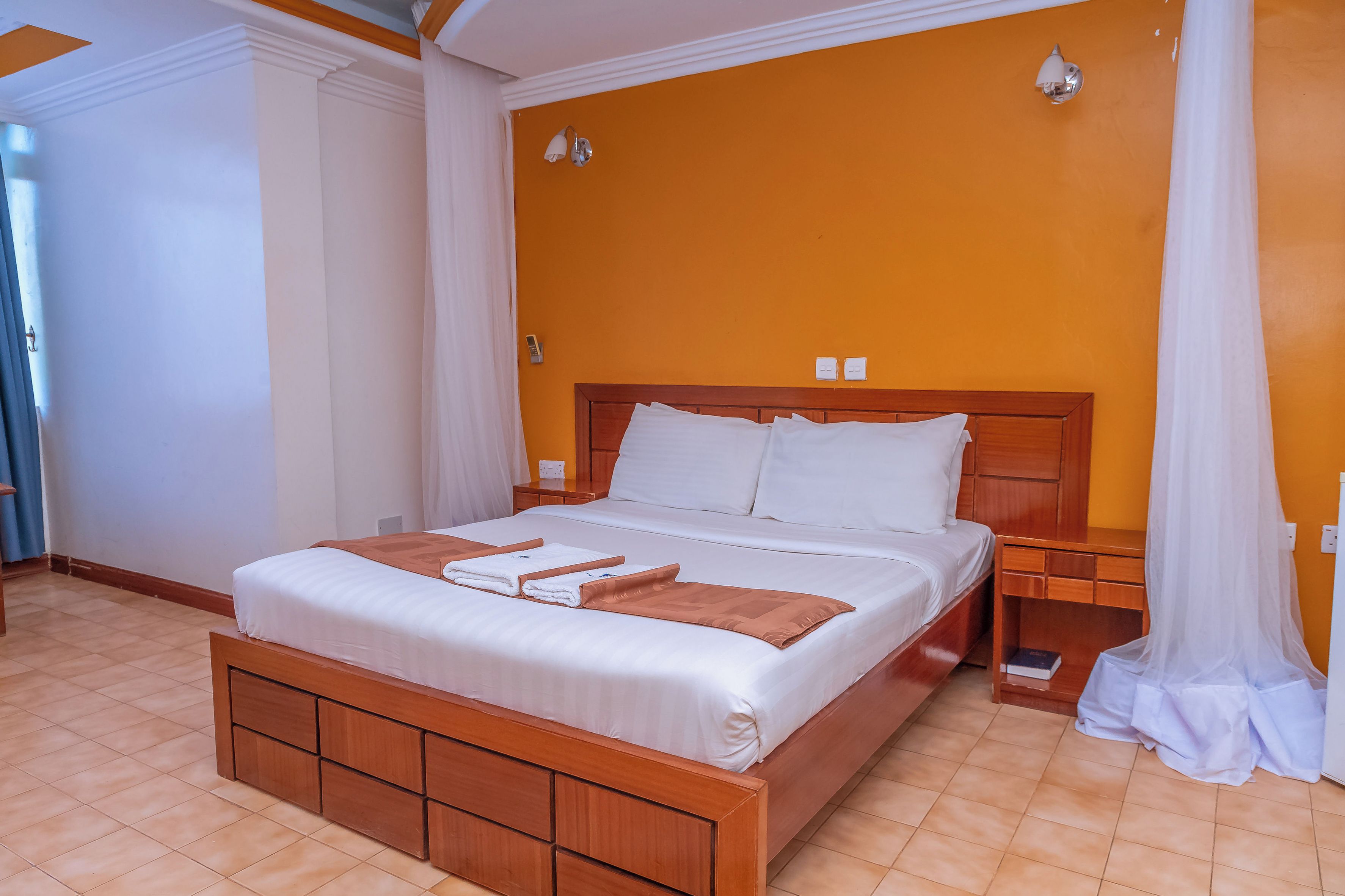 A hotel room with a large bed, wooden headboard, white linens, and a bright orange accent wall.