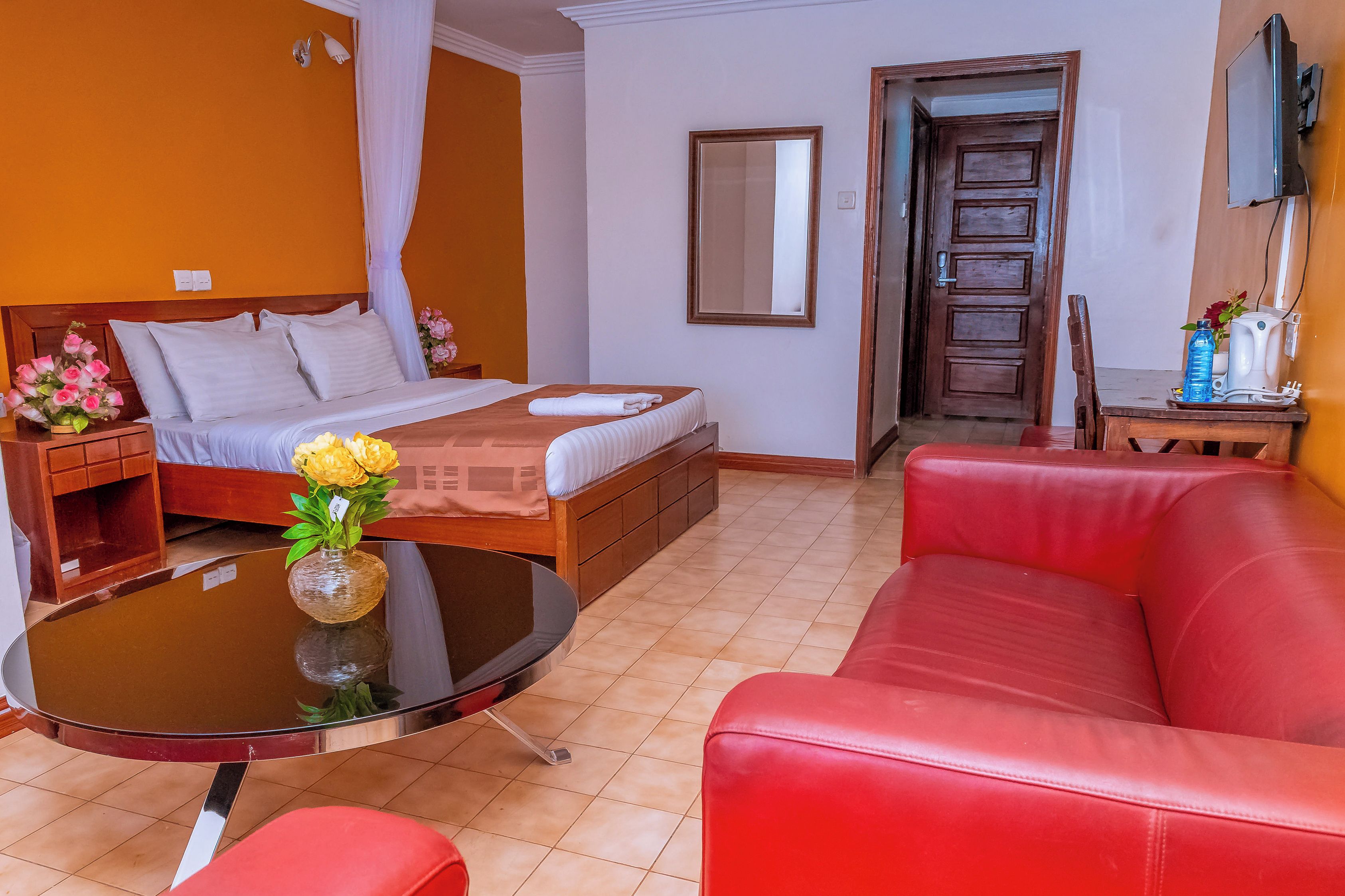 Una habitación de hotel con una cama, un sofá de cuero rojo, una mesita con un florero, un espejo y una puerta que da a otra habitación.
