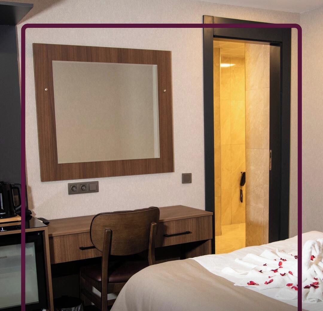 Een hotelkamer met een houten bureau, een stoel, een bed met wit beddengoed, een grote spiegel en een deur naar een badkamer.