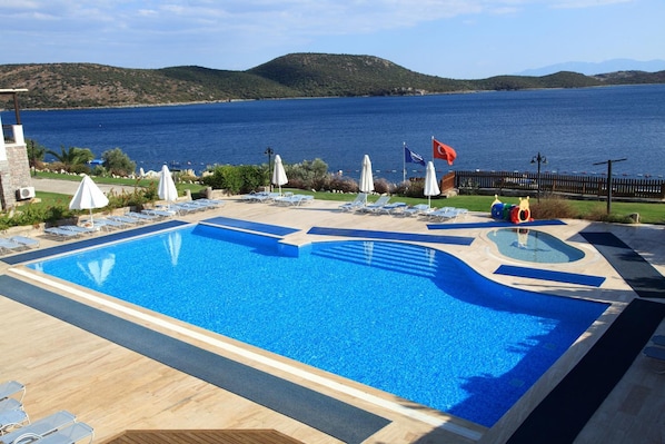 A swimming pool with lounge chairs and umbrellas, surrounded by a wooden deck and a view of the sea and hills. - Aktur Residence (Bodrum)