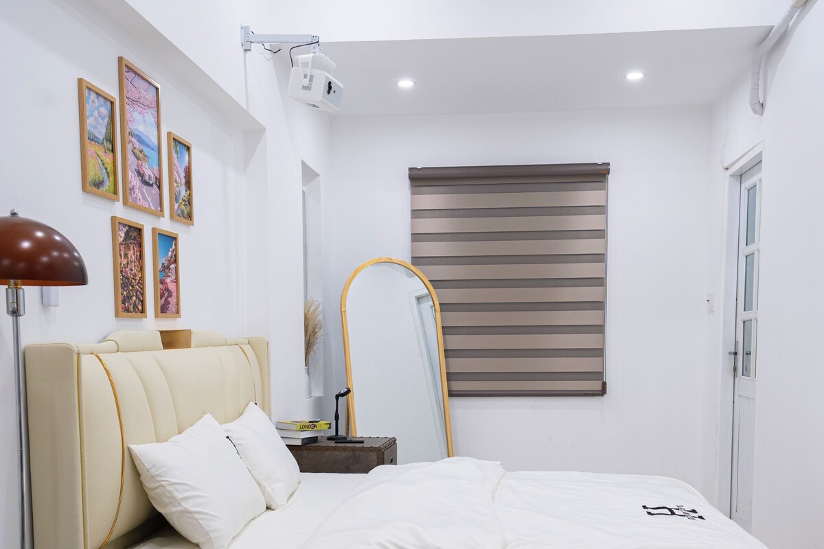 Quarto com cama, mesa de cabeceira, abajur e janela com persianas.