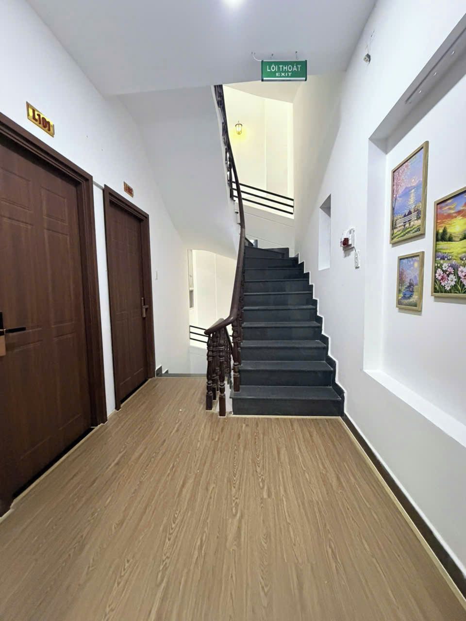 Un pasillo con una escalera que conduce a una salida, suelo de madera y cuadros en las paredes.