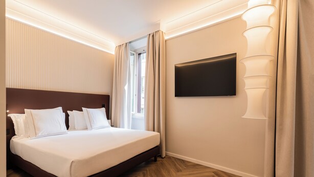 A hotel room with a large bed, a flat-screen TV mounted on the wall, and a window with curtains.